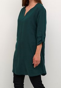 Teal tunika med v-hals, trekvart lange opsmøgede ærmer og en løs pasform. Fremstillet af et blødt, struktureret stof. Subtile sideslidsdetaljer.