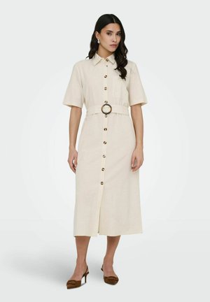 Žena oblečená v béžových midi šatách na gombíky s krátkymi rukávmi, s okrúhlou prackou na opasku a hnedými lodičkami na podpätku, stojaca pred jednoduchým pozadím.