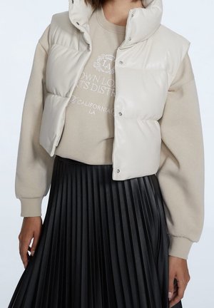 Femme portant une veste sans manches matelassée beige par-dessus un sweat-shirt beige et une jupe plissée noire, debout devant un fond uni.