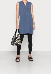 Blå ärmlös tunika med krage och knappar, ihop med svarta leggings och stiliga svarta sandaler, hållandes en vävd svartvit väska.