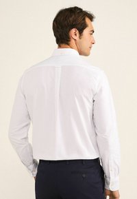 Camisa blanca de manga larga con un pliegue en la parte trasera, puños abotonados y cuello con solapa. La tela parece suave y ligera.