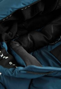 Intérieur d'une veste doudoune bleue avec doublure en polaire noire, tissu lisse et plusieurs compartiments. Présente des coutures et des étiquettes contrastantes.