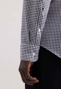 Geruite shirt met een wit en marineblauw patroon, katoenen stof en knoopmanchetten. Hand in beeld met een donkere huidskleur.