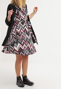 Klänning med zickzacksmönster i rött, svart och vitt, med en utsvängd kjol och korta ärmar, tillsammans med en svart kofta och ankelstövlar.