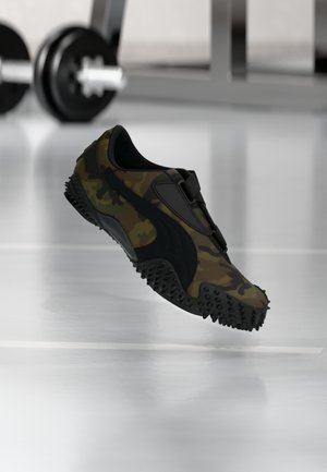 Zapatilla deportiva con estampado de camuflaje y suela texturizada, flotando sobre el suelo del gimnasio, con pesas y equipo de entrenamiento desenfocados al fondo.
