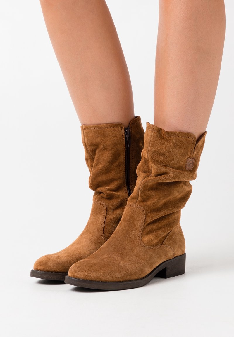 Bottes chevilles en daim marron avec design froncé, fermeture éclair latérale et talon plat. Caractérisées par un embout rond et des détails subtils cousus sur les côtés.