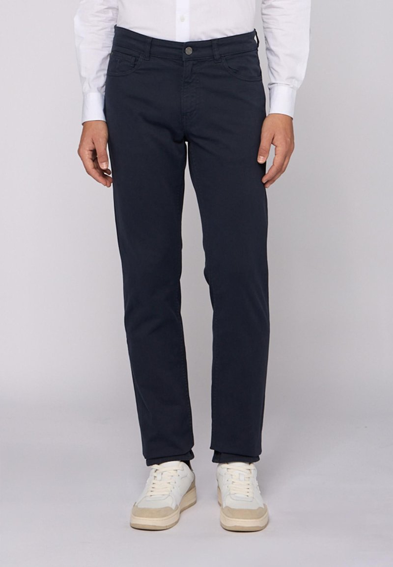 Pantalones azul marino fabricados en una mezcla de algodón, con un ajuste slim, diseño de cinco bolsillos y cierre con botón. Se llevan con zapatillas blancas.