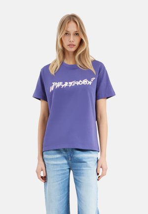 T-shirt viola con una vestibilità comoda e maniche corte. Presenta una scritta grafica bianca sul petto. Materiale in cotone morbido. Indossato con jeans blu chiaro.