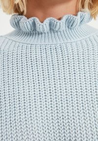 Jasnoniebieski sweter z dzianiny z falbaną przy wysokim dekolcie, pokazany na osobie z krótkimi blond włosami.