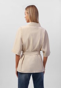 Blusa beige avvolgente con maniche corte, cintura con cinturino e tessuto testurizzato. Abbinata a pantaloni di jeans blu, vista da dietro.
