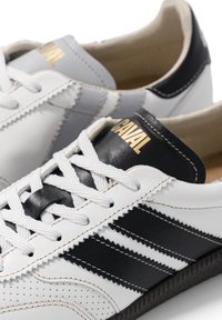 Une paire de baskets en cuir blanches avec des bandes noires, des lacets blancs et le mot "CAVAL" en or sur la languette.