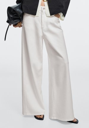 Personne portant un pantalon blanc à jambes larges, des chaussures noires à bout ouvert, tenant un sac noir dans une main et portant une veste noire par-dessus une chemise claire.