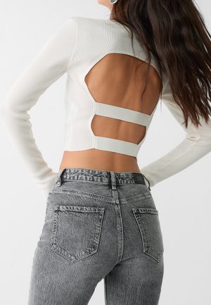 Femme portant un crop top blanc à manches longues côtelé avec un dos ouvert et deux brides horizontales, associé à un jean en denim gris taille haute.