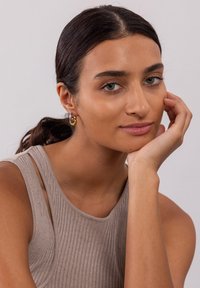 Rippentop in Beige, helle Hautfarbe und lange dunkle Haare. Trägt goldene und silberne Creolen. Glatte Hauttextur und neutraler Hintergrund.