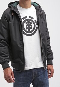 Czarna kurtka na zamek z kapturem, z prążkowanymi mankietami i zielonym, kraciastym podszyciem. Noszona na białej koszulce z dużym czarnym logo graficznym.