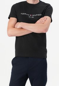 Mustapaita, jossa on lyhyet hihat, ja etupuolella valkoinen ja punainen logo-teksti "TOMMY HILFIGER EST. 1985". Tasainen pinta.