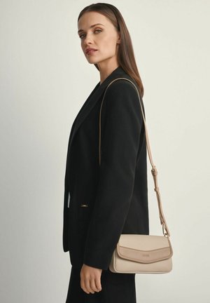 Kobieta z długimi brązowymi włosami, w czarnym blezerze, nosząca beżową skórzaną torbę na ramię, na tle jednolitego jasnego tła.