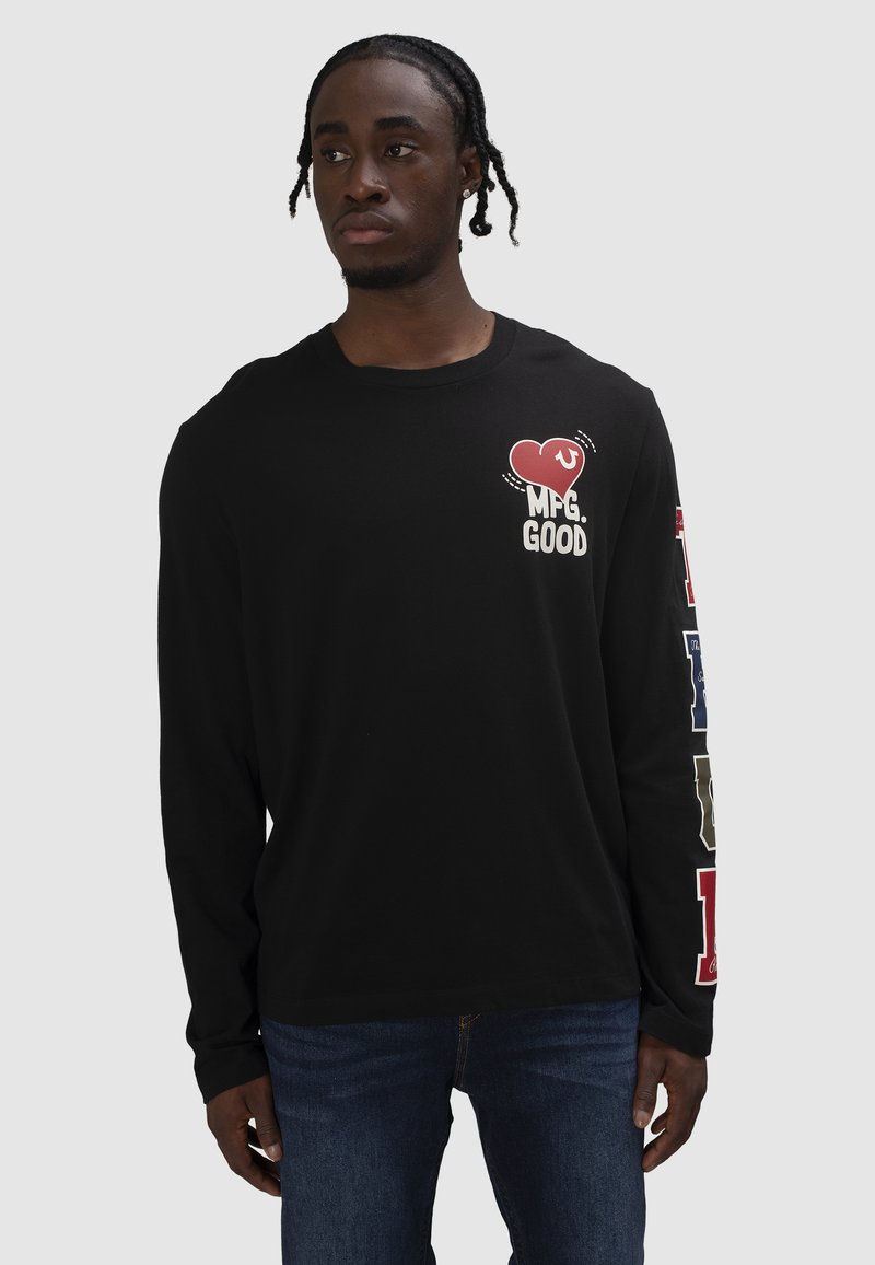 Mann mit geflochtenem Haar, der ein schwarzes Langarmshirt mit rotem Herz und dem Schriftzug "MFG. GOOD" auf der Brust sowie grafischen Buchstaben am rechten Ärmel trägt.