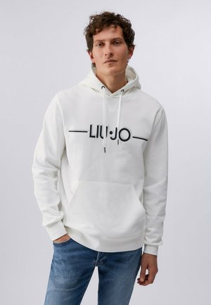 Junger Mann mit lockigem Haar, der einen weißen Hoodie mit dem "LIU JO"-Logo und blaue Jeans trägt, steht vor einem einfarbigen hellgrauen Hintergrund.
