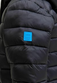 Svart puffjacka med quiltade sektioner och en blå rektangulär etikett med "JACK & JONES" och "ETABLERAD ÅR 1990" präglad.