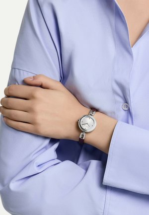 Montre argentée avec un cadran rond, un cadran texturé argenté et un bracelet en cuir violet. Portée sur la manche d'une chemise bleu clair, mettant en avant son design moderne.