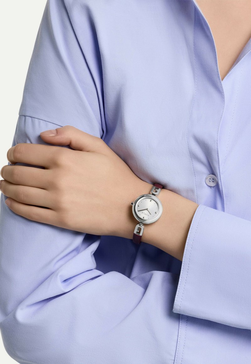 Montre argentée avec un cadran rond, un cadran texturé argenté et un bracelet en cuir violet. Portée sur la manche d'une chemise bleu clair, mettant en avant son design moderne.