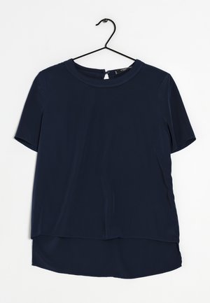Top blu navy a maniche corte realizzato in tessuto morbido, con scollatura rotonda e orlo posteriore leggermente più lungo, appeso a un gancio nero.