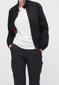 Schwarze Bomberjacke aus glattem Material mit hohem Kragen, Frontreißverschluss und rotem Karofutter; kombiniert mit einem weißen T-Shirt und schwarzen Cargo-Hosen.