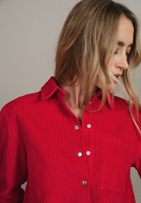 Camicia di velluto a coste rossa con colletto, caratterizzata da una texture a coste verticali, bottoni argentati e una tasca a toppa sul lato sinistro.