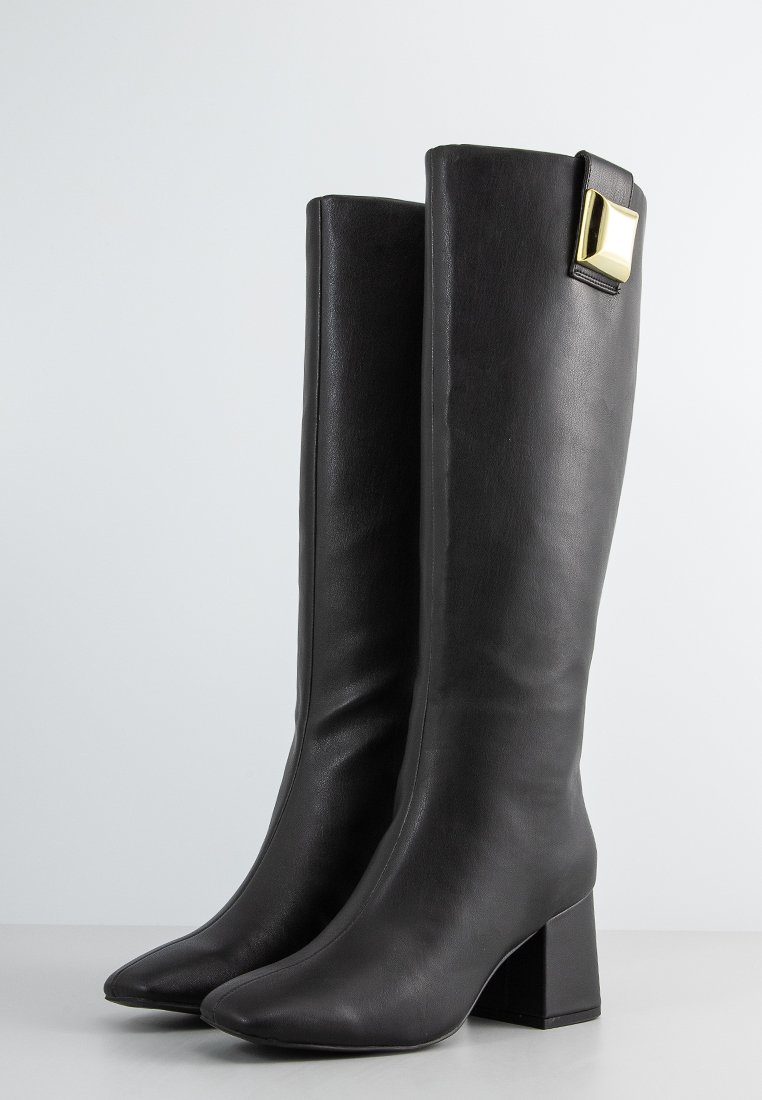 Schwarze knielange Lederstiefel mit Blockabsätzen und einer goldenen quadratischen Schnalle oben auf einem schlichten weißen Hintergrund.