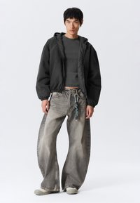 Jonge man in een zwart hoodie-jack, donkergrijze cropped shirt, brede vervaagde grijze jeans met sleutelhanger, en lichtgrijze schoenen, staande tegen een witte achtergrond.
