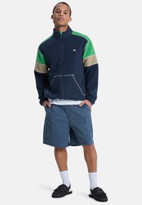 Ναυτική ζακέτα από φλις με πράσινες και μπεζ λεπτομέρειες, κλείσιμο με φερμουάρ και τσέπες μπροστά, σε συνδυασμό με μπλε cargo σορτς και μαύρες σαγιονάρες.