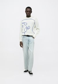 Witte gebreide trui met een blauw abstract gezicht ontwerp, gecombineerd met lichtblauwe spijkerbroek en zwarte sneakers. Model staat tegen een effen achtergrond.