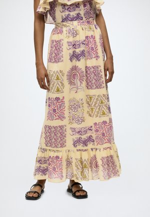 Jupe maxi jaune fluide avec motifs patchwork floraux et géométriques violets et roses, portée avec des sandales plates noires à brides.