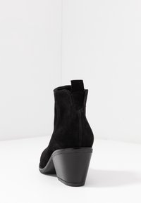 Botas de tobillo de ante negro con punta redondeada, tacón de cuña apilada y una tira para tirar. La parte superior tiene sutiles acentos de costura.