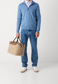 Pull bleu clair avec une fermeture éclair, jean bleu et baskets blanches ; tenant un sac en daim beige avec des accents en cuir marron.