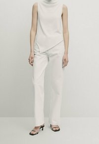 White sleeveless top with high neck, asymmetrical hem; paired with white straight-leg trousers and black open-toe heels.