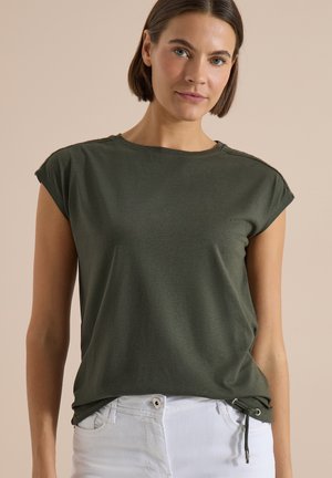 Ung kvinde iført en olivengrøn top med ærmer og hvide bukser, stående mod en ensfarvet beige baggrund.