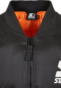 Giacca bomber nera con colletto alto, foderatura arancione, zip frontale e grande logo bianco sul lato sinistro. Tessuto dalla texture liscia.