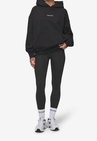 Felpa nera con tasca frontale e logo, abbinata a leggins neri. Scarpe sportive bianche con dettagli neri e calzini bianchi con logo.