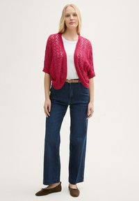 Cardigan léger en tricot ouvert de couleur rose vif avec des manches courtes, associé à un t-shirt blanc, un jean foncé taille haute et des ballerines marron.