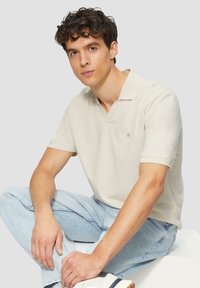 Beige poloshirt met een korte kraag, gemaakt van textuurstof. Gecombineerd met lichtblauwe jeans en witte sneakers met marineblauwe accenten.