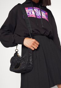 Černá prošívaná kabelka s zakřiveným tvarem, se zlatými kovovými detaily a logem. Doplněna černou plisovanou sukní a mikinou.
