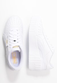 Baskets blanches avec une tige en cuir lisse, des détails perforés, des lacets plats blancs et une semelle épaisse texturée. Accent de logo doré visible.