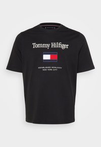 Must musta t-särk, millel on "Tommy Hilfiger" tekst, ristkülikukujuline logo punase, valge ja sinisega ning tekst "Established 1985 New York City."