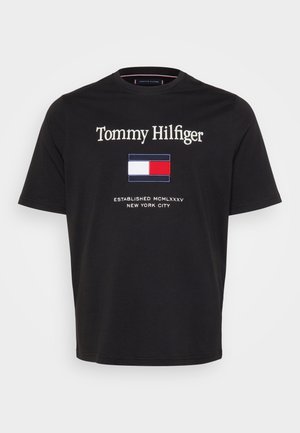 Melnā kokvilnas T-kreklā ir redzams "Tommy Hilfiger" teksts, taisnstūrveida logo ar sarkanu, baltu un zilu krāsu, kā arī teksts "Iznākusi 1985. gadā Ņujorkā".