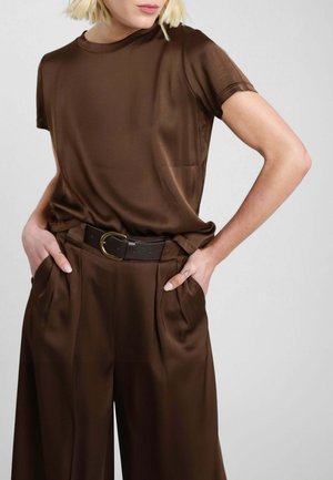 Donna che indossa un top marrone in raso a maniche corte e pantaloni larghi abbinati con una cintura nera, con le mani nelle tasche, su uno sfondo semplice.