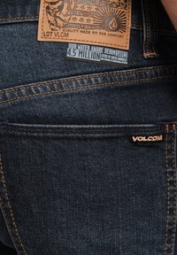 Vaqueros de mezclilla azul oscuro con costuras naranjas. Presentan un parche de cuero en la parte trasera con un diseño embossado y una pequeña etiqueta de marca.