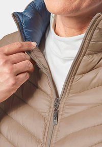 Giacca imbottita marrone chiaro con colletto zip. Il colletto interno è blu, con una texture trapuntata e una superficie liscia.
