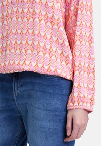 Blouse à manches longues avec un motif abstrait rose et orange, fabriquée en tissu doux avec des poignets élastiques, assortie à un jean bleu.